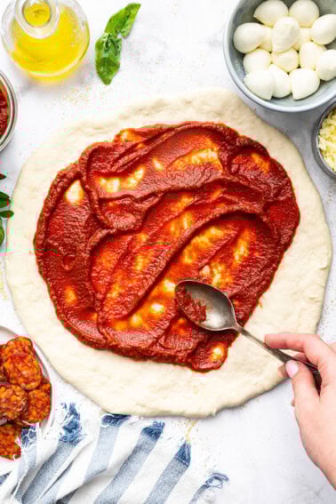 Spreading homemade pizza sauce over pizza dough with a spoon, ready for toppings.