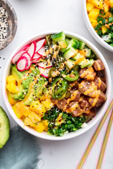 Finished poke bowl with all toppings - ahi tuna, jalapenos, cucumbers, radishes, avocado, mango, seaweed salad and scallions. Bowl is drizzled with sriracha mayo and topped with sesame seeds.