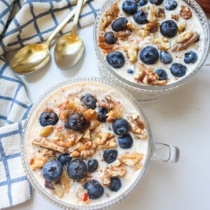 Two glass bowls of overnight oats topped with fresh blueberries and walnuts, set on a white table with gold spoons and a blue-and-white napkin.