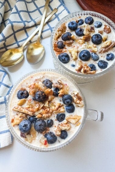 Two glass bowls of overnight oats topped with fresh blueberries and walnuts, set on a white table with gold spoons and a blue-and-white napkin.