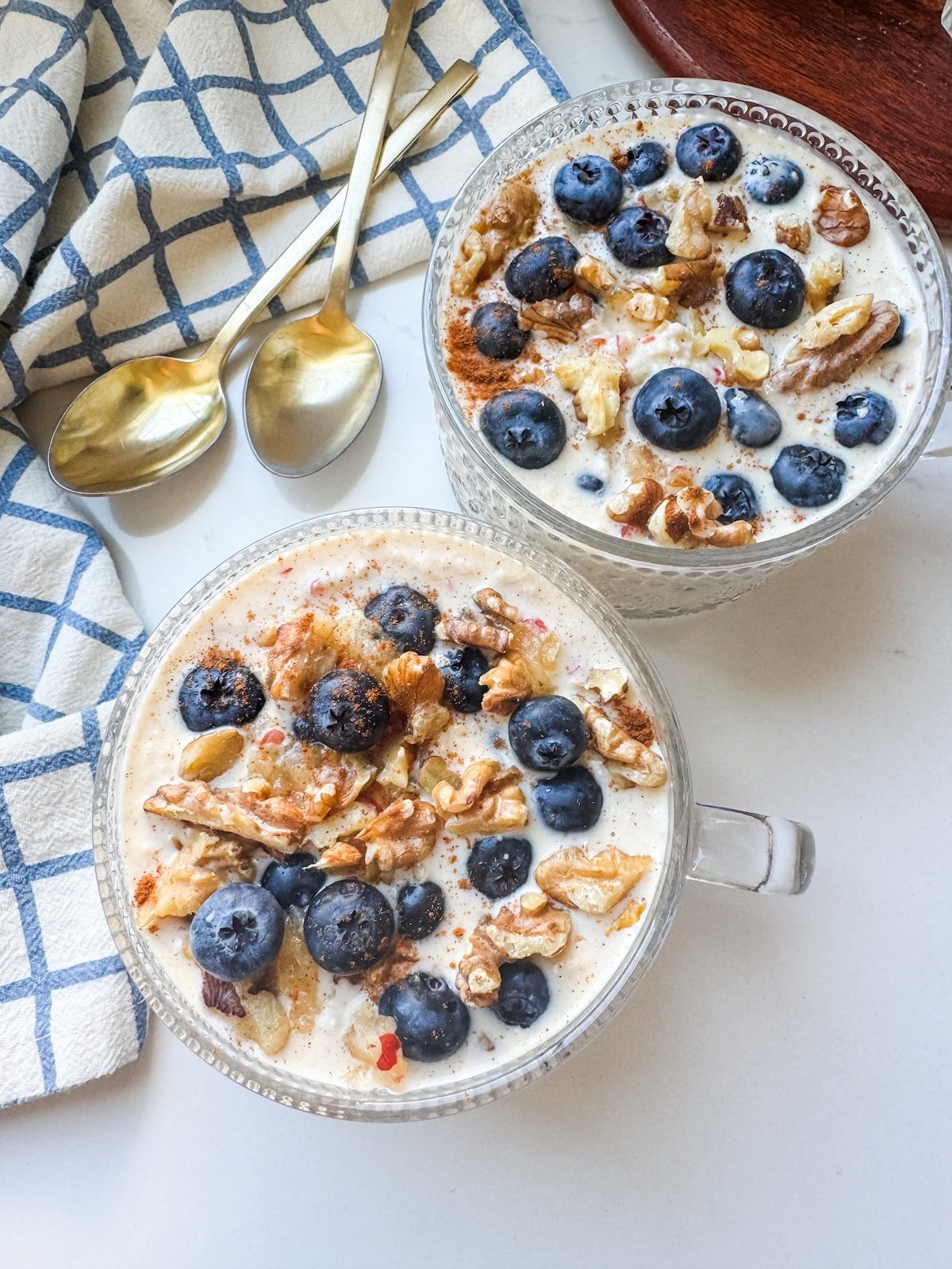 Two glass bowls of overnight oats topped with fresh blueberries and walnuts, set on a white table with gold spoons and a blue-and-white napkin.