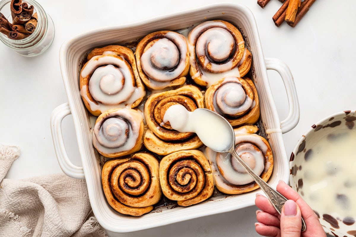 A woman's hand drizzling the glaze over the 9 protein cinnamon rolls in a white square baking dish.