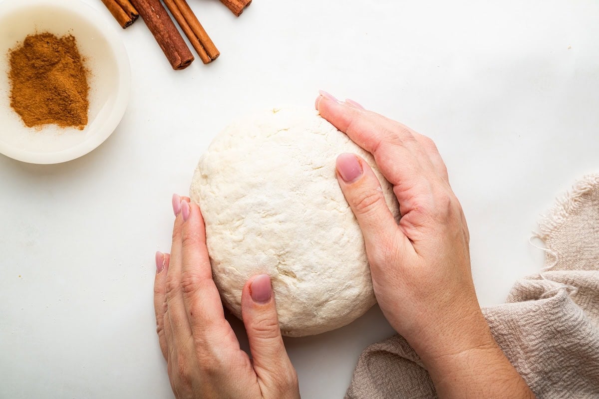 A woman's hands forming a ball of protein cinnamon roll dough. There are cinnamon sticks and ground cinnamon in the background.