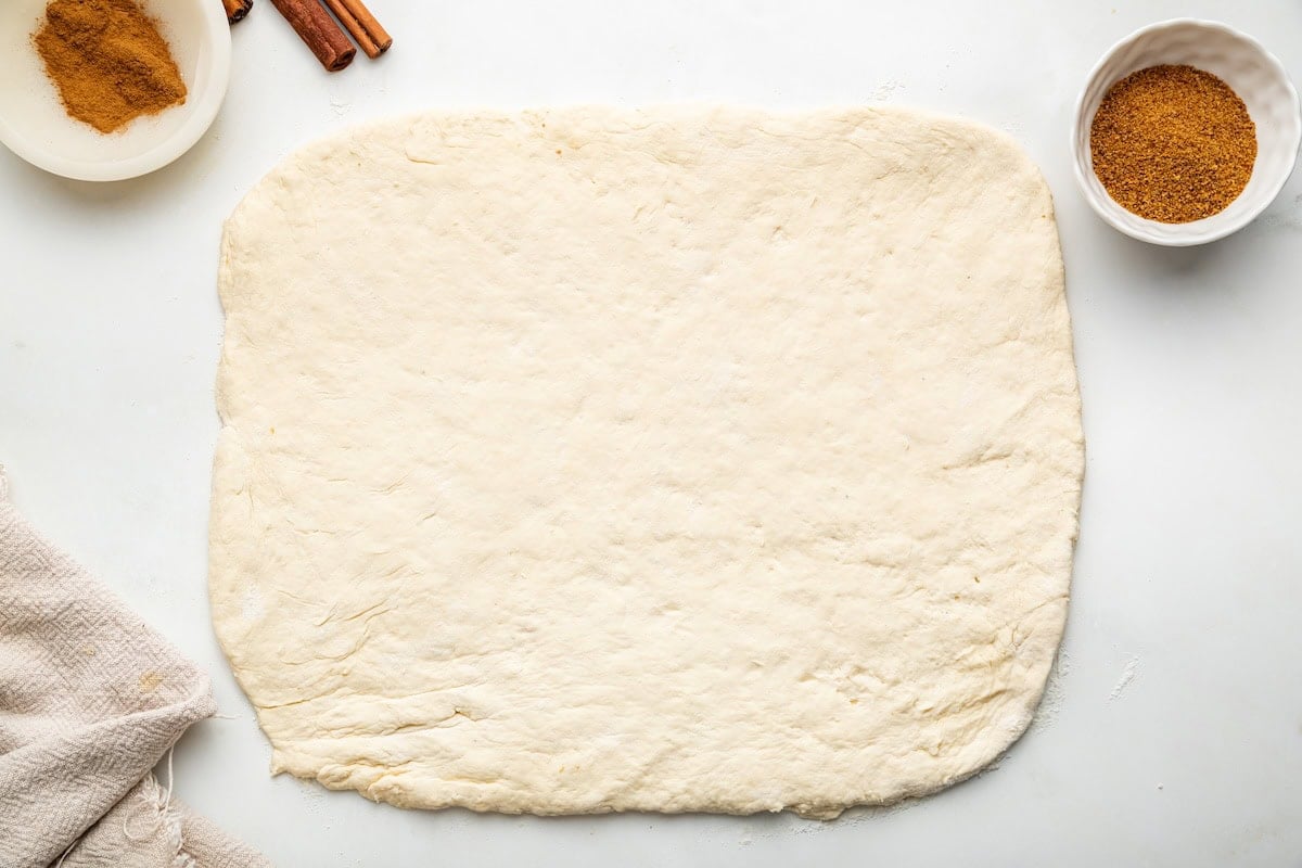 The dough for the protein cinnamon rolls rolled out in a rectangular shape on a counter. Coconut sugar and ground cinnamon are in the background.