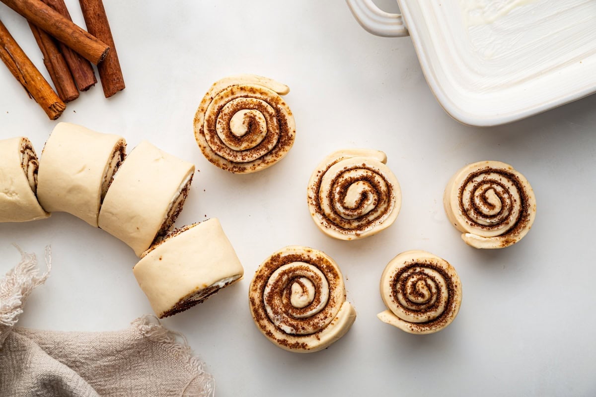 9 protein cinnamon rolls cut out on a counter. A white baking dish and cinnamon sticks are in the background.