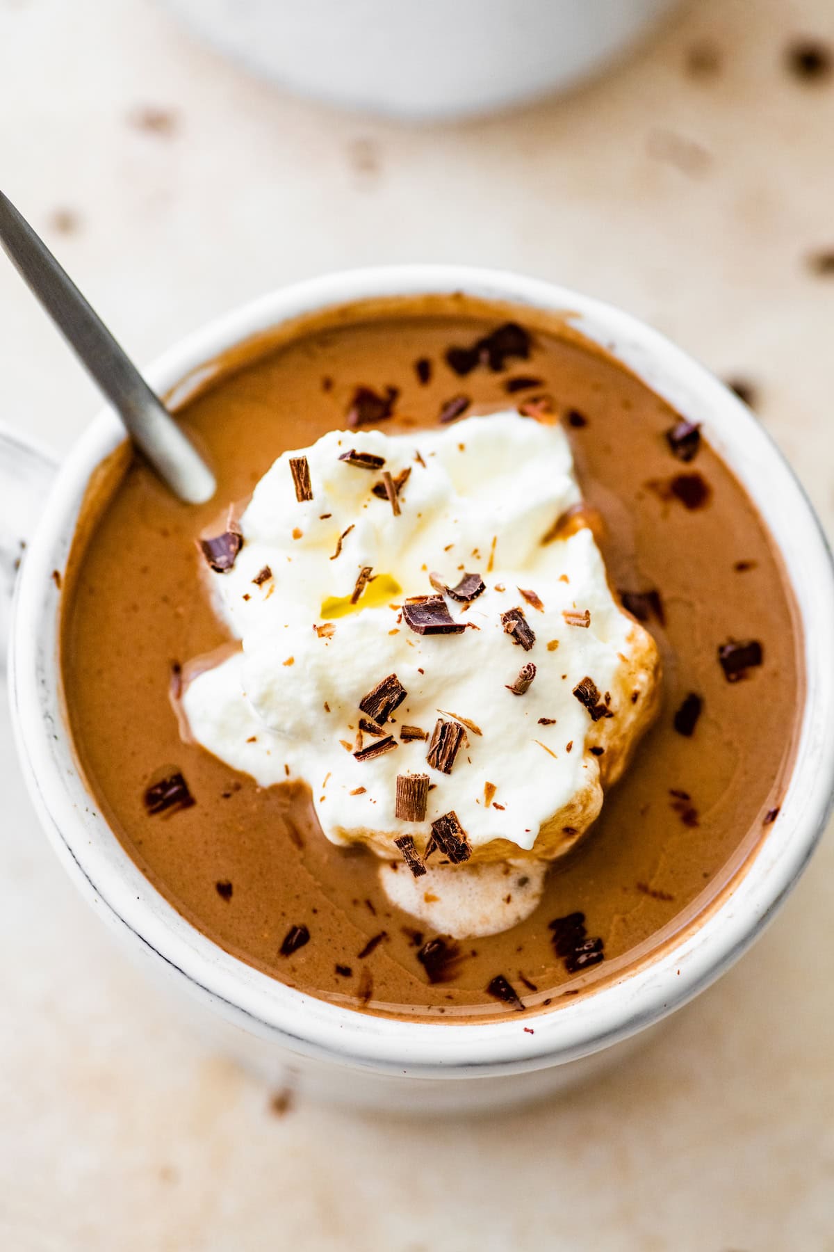 Protein hot chocolate in a white ceramic mug topped with whip cream and shaved chocolate. A silver spoon is in the mug.