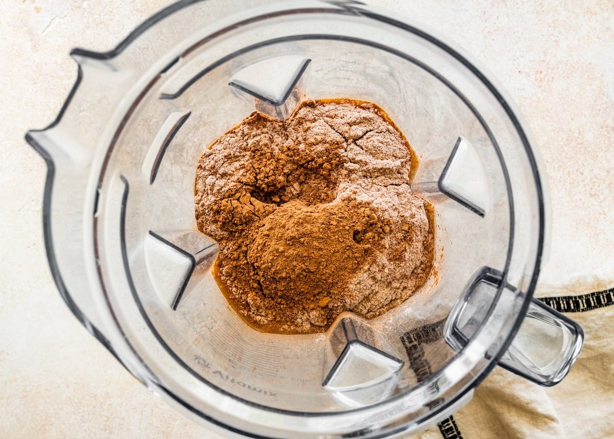 The ingredients for the protein hot chocolate (chocolate protein powder, cocoa powder, maple syrup, milk, vanilla and salt) in the base of a Vitamix blender