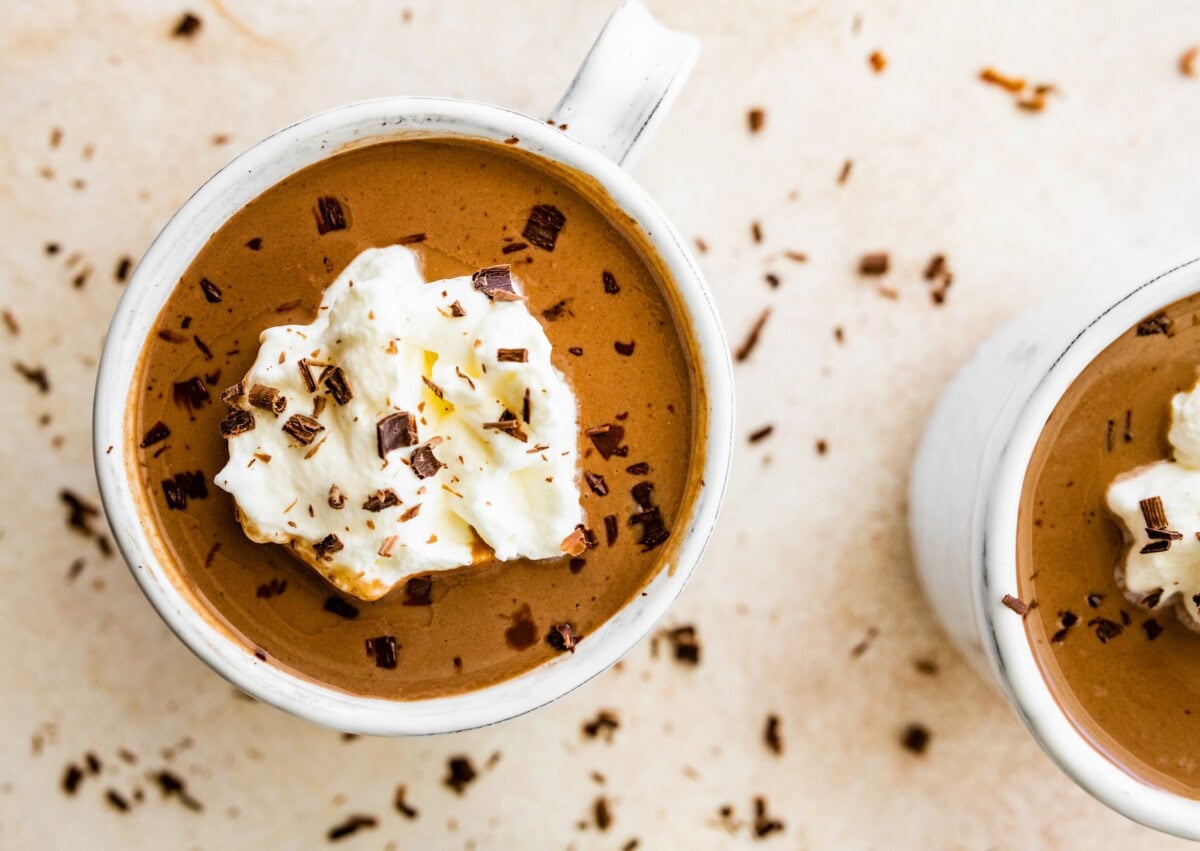 Two white ceramic mugs filled with the protein hot chocolate. Both are topped with whip cream and shaved chocolate.