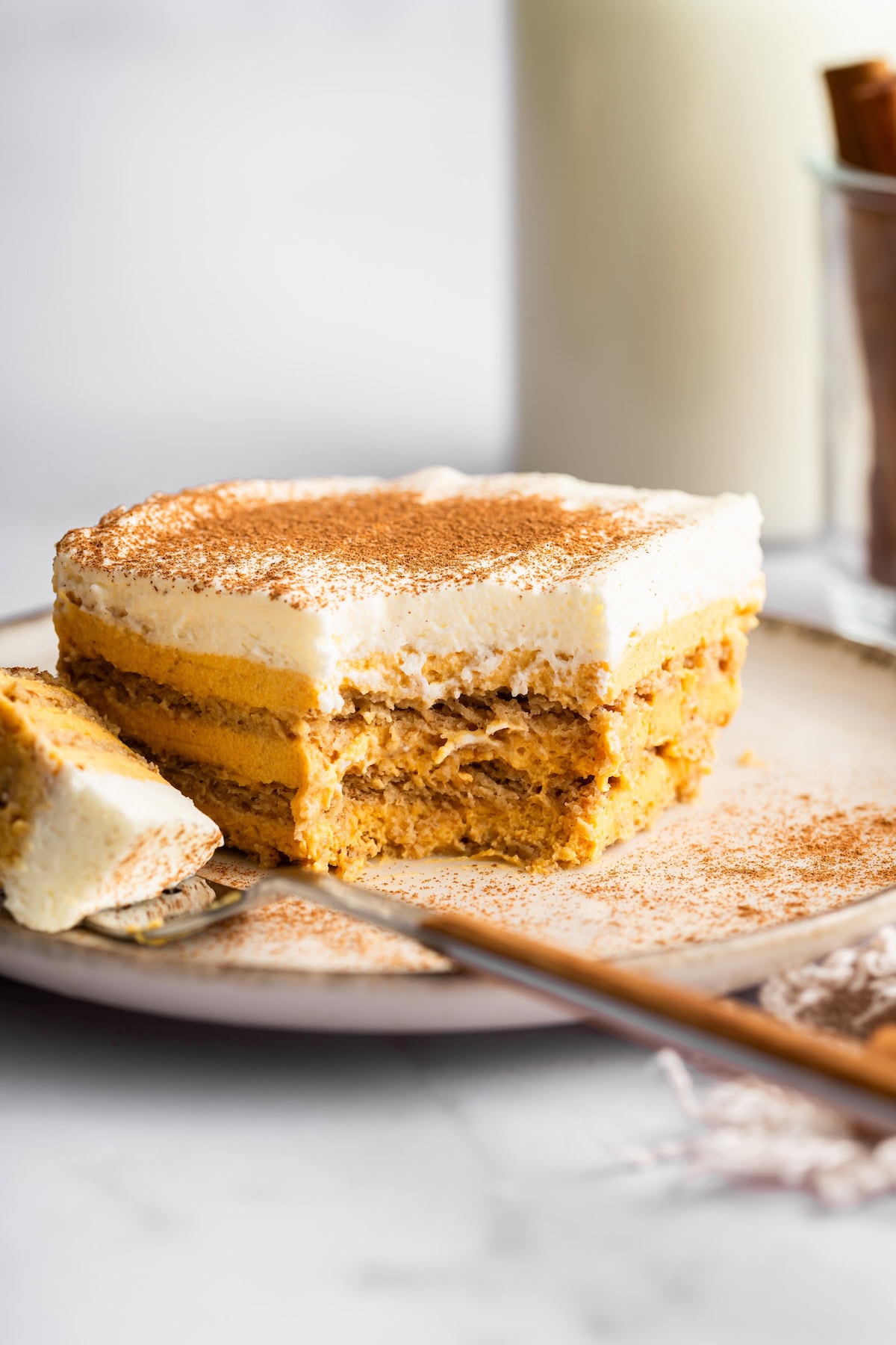A slice of pumpkin icebox cake topped with cinnamon on a plate, with a bite taken out to show creamy layers of pumpkin and whipped topping.