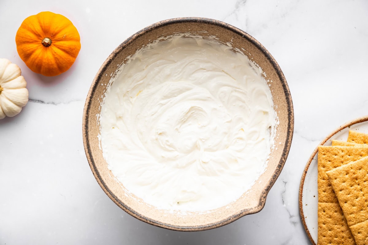 A mixing bowl filled with a smooth white whipped cream, next to graham crackers and small pumpkins.