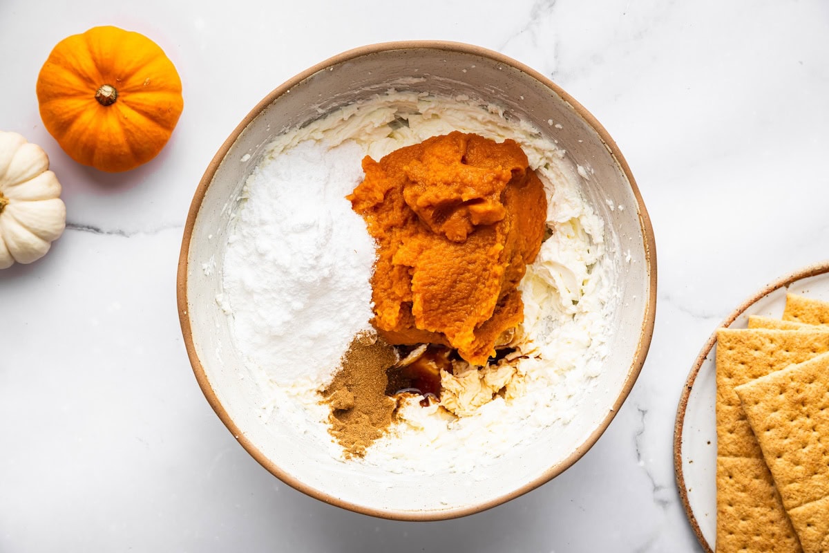 A mixing bowl containing pumpkin puree, powdered sugar, spices, and whipped cream before mixing, with graham crackers and pumpkins beside it.