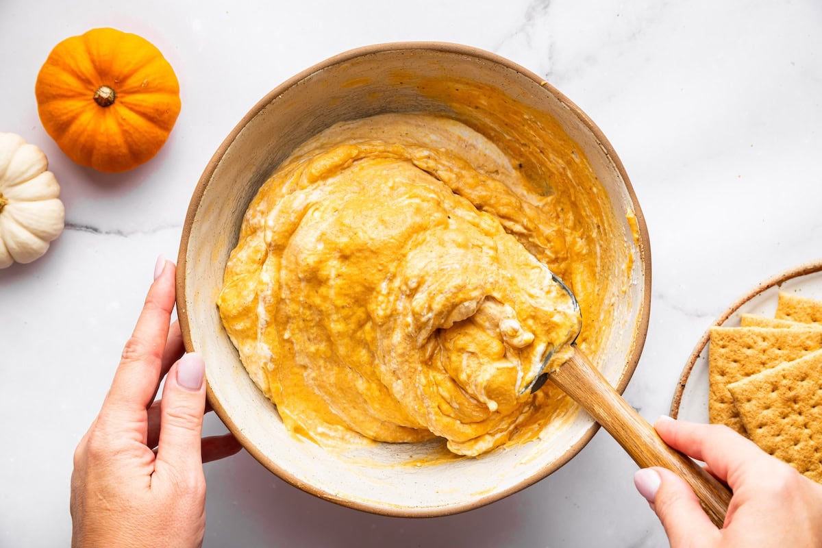 A bowl of pumpkin mixture being folded together with a spatula, surrounded by graham crackers and small pumpkins.