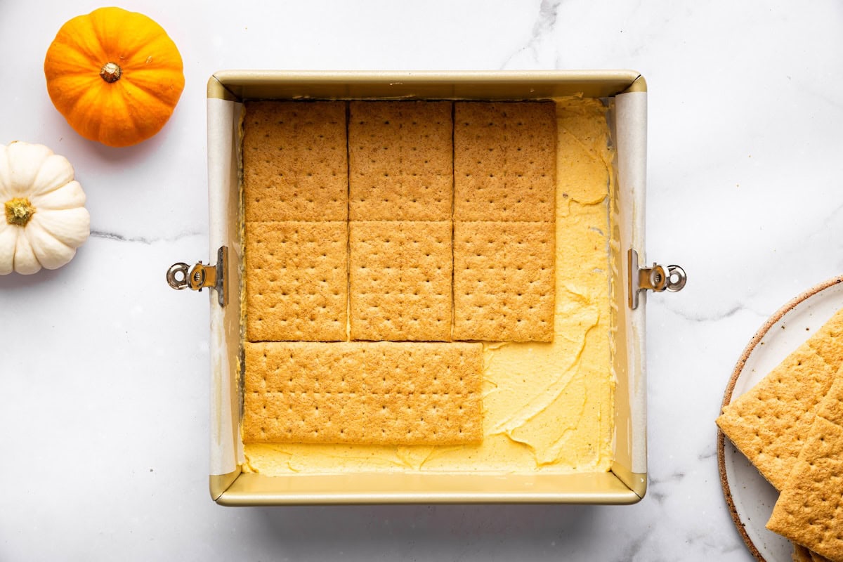 A baking pan filled with pumpkin filling topped with a single layer of graham crackers, with more crackers and mini pumpkins nearby.