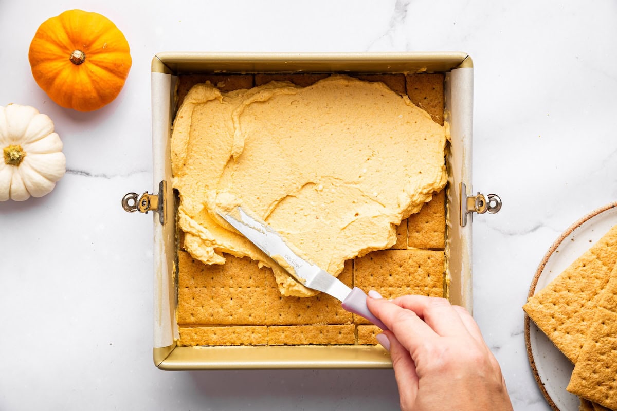 A hand spreading pumpkin filling over a layer of graham crackers in a baking pan with mini pumpkins and extra graham crackers to the side.