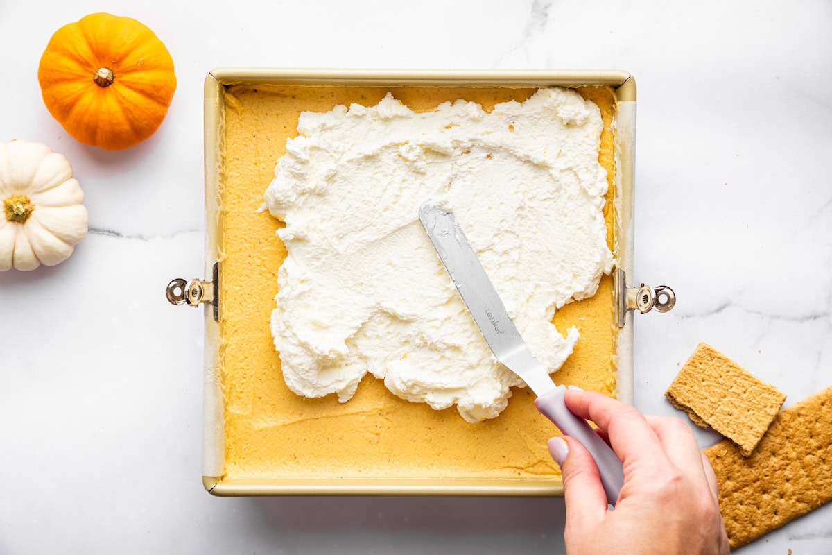 A hand spreading a layer of whipped topping over pumpkin filling in a square baking pan with mini pumpkins and graham crackers nearby.