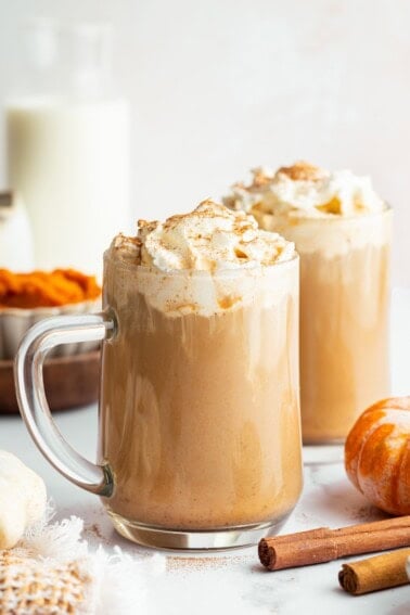 Two glass mugs filled with pumpkin spice lattes topped with whipped cream and cinnamon, set among pumpkins, milk, and fall spices.