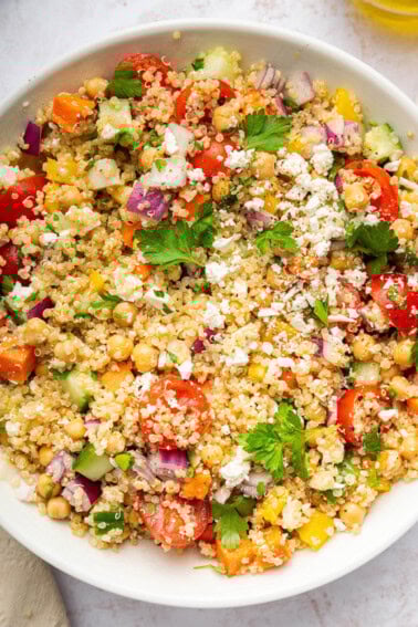 A mixed quinoa chickpea salad with tomatoes, cucumbers, feta, and parsley, in a white bowl.