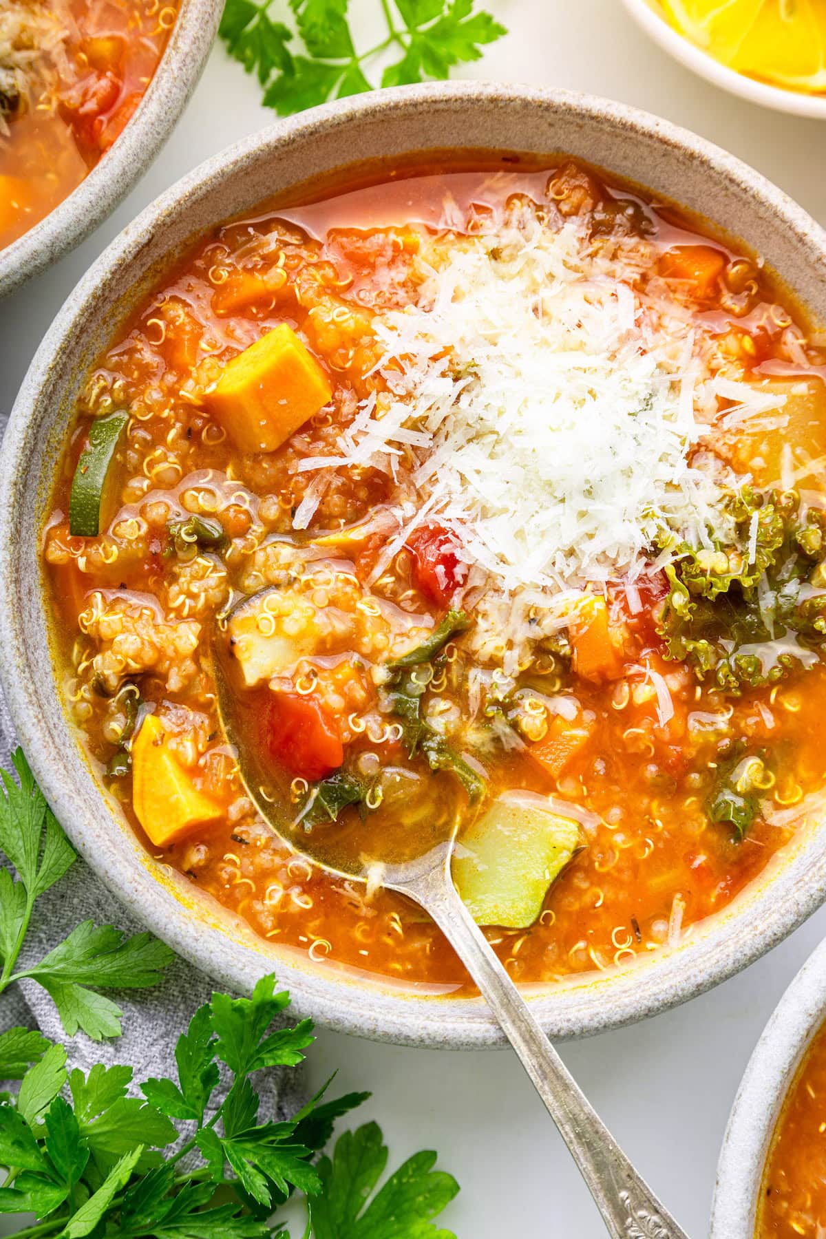A full bowl of quinoa vegetable soup garnished with fresh Parmesan and a metal spoon is in the soup.
