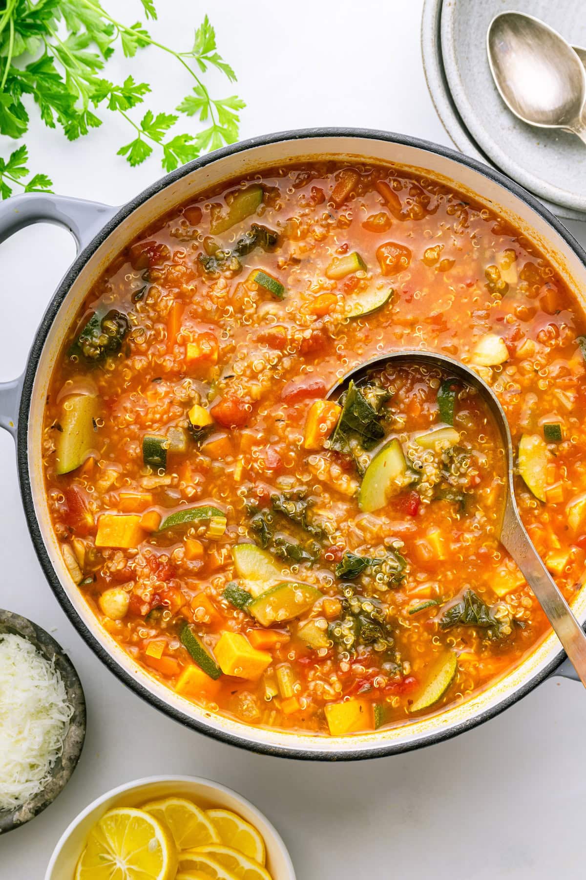 A large pot of vegetable quinoa soup with a metal serving spoon in the pot. The soup is loaded with zucchini slices, fresh kale, vegetable broth, sauteed vegetables, diced sweet potato, and tomatoes.
