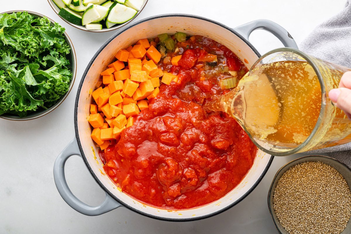 A womans hand pours vegetable broth into a large pot filled with sauteed vegetables, diced sweet potato, and tomatoes.