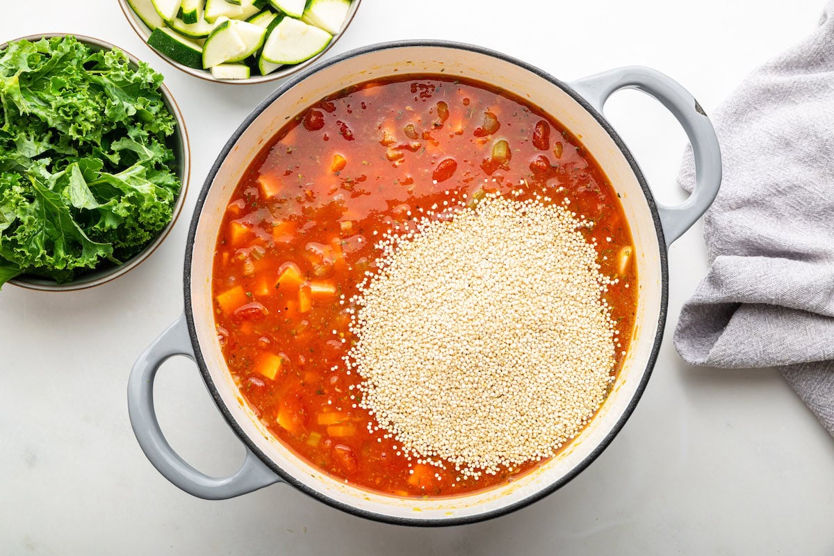Quinoa is added to a large pot with vegetable broth, sauteed vegetables, diced sweet potato, and tomatoes.