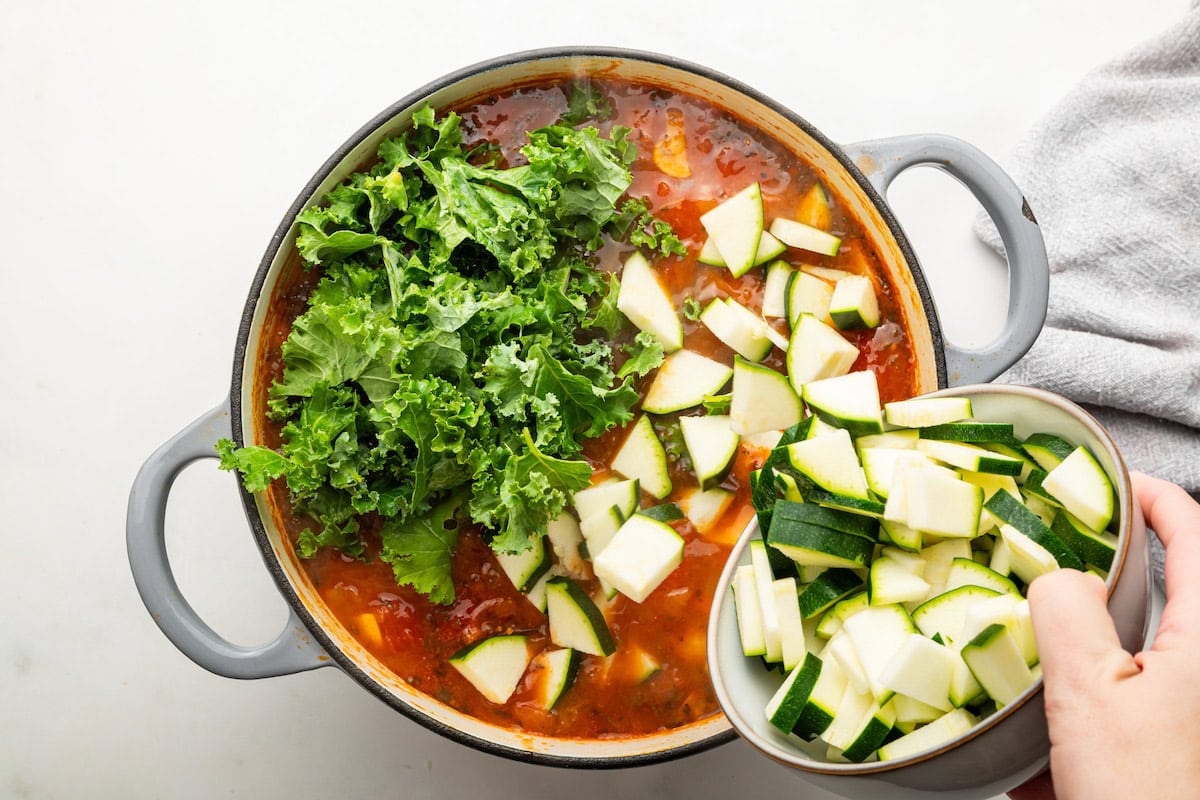 Zucchini slices and fresh kale are added to a large pot of quinoa, vegetable broth, sauteed vegetables, diced sweet potato, and tomatoes.