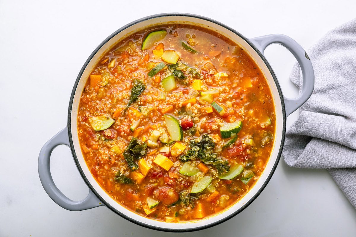 A large pot of vegetable quinoa soup with zucchini slices, fresh kale, vegetable broth, sauteed vegetables, diced sweet potato, and tomatoes.