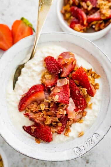 Cottage cheese in a bowl topped with roasted strawberries, rolled oats and coconut. There is a silver spoon in the bowl.