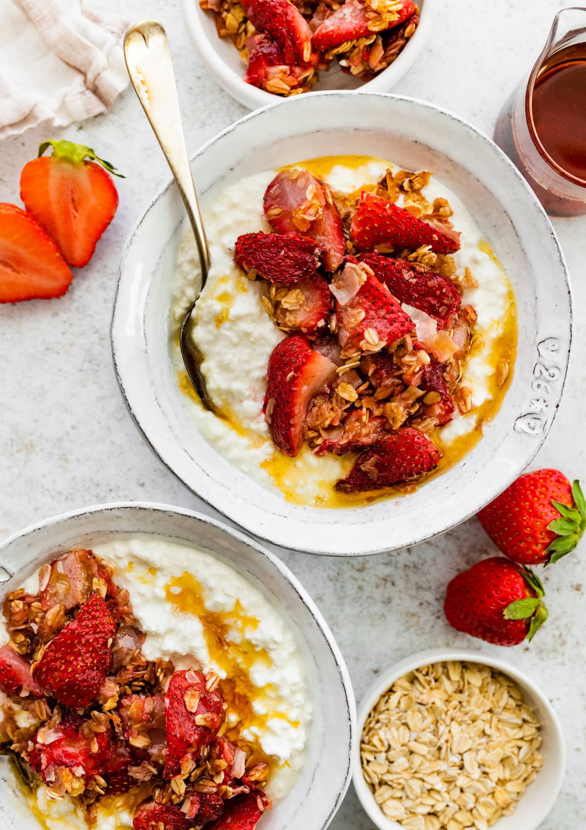 Two bowls filled with cottage cheese that's topped with roasted strawberries, rolled oats, coconut and a drizzle of maple syrup.