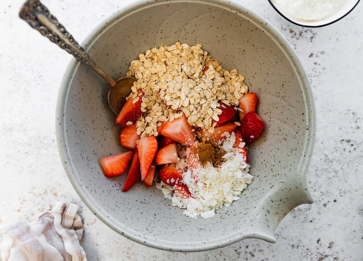 Rolled oats, coconut, chopped fresh strawberries and cinnamon in a mixing bowl with a silver spoon.