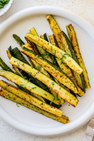 Golden roasted zucchini sticks on a white plate, garnished with chopped parsley.