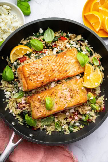 Two salmon filets in a skillet served over a bed of orzo combined with roasted red peppers, chopped dates, feta and swiss chard. It's topped with lemon slices and fresh basil.