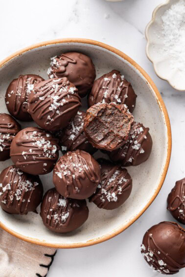 Bowl of chocolate protein balls stacked with flaky sea salt on top and one bitten into.