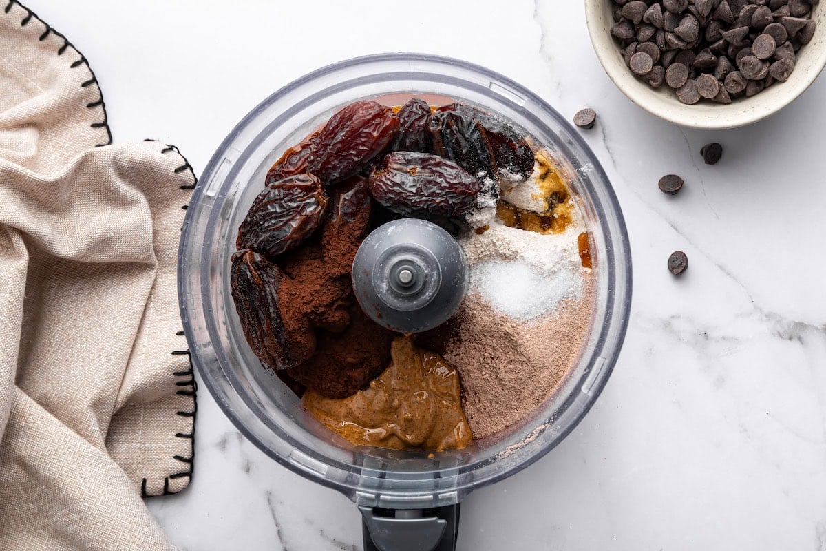 Food processor filled with dates, nut butter, cacao, and protein powder before blending.