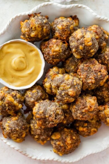 Baked sausage balls piled on a plate beside a dish of creamy mustard sauce.