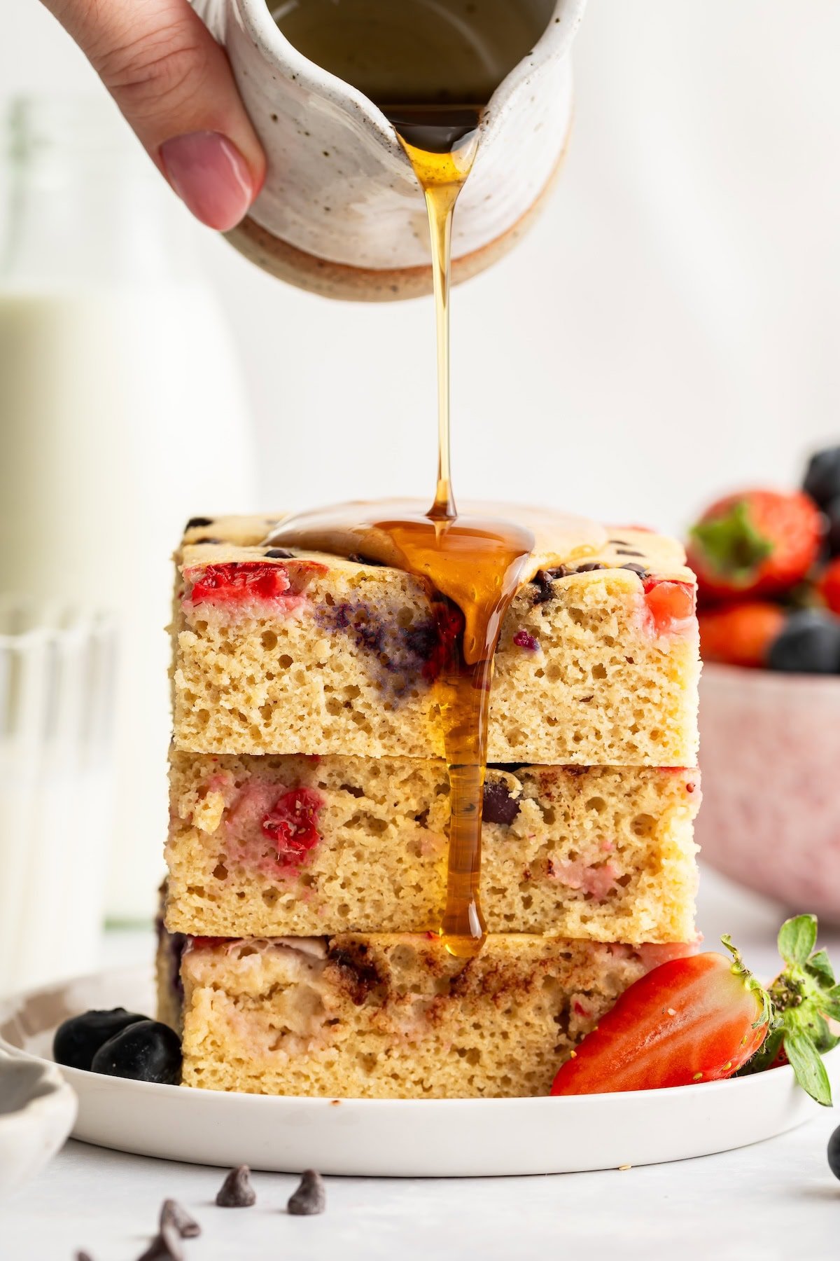 Stack of fluffy sheet pan pancakes topped with strawberries and syrup being poured.