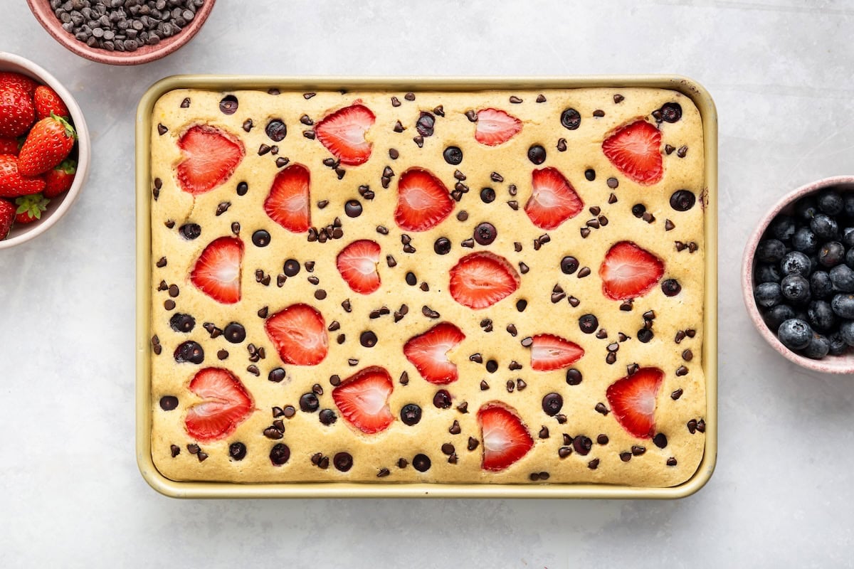 Sheet pan pancakes topped with sliced strawberries, blueberries, and chocolate chips after baking.