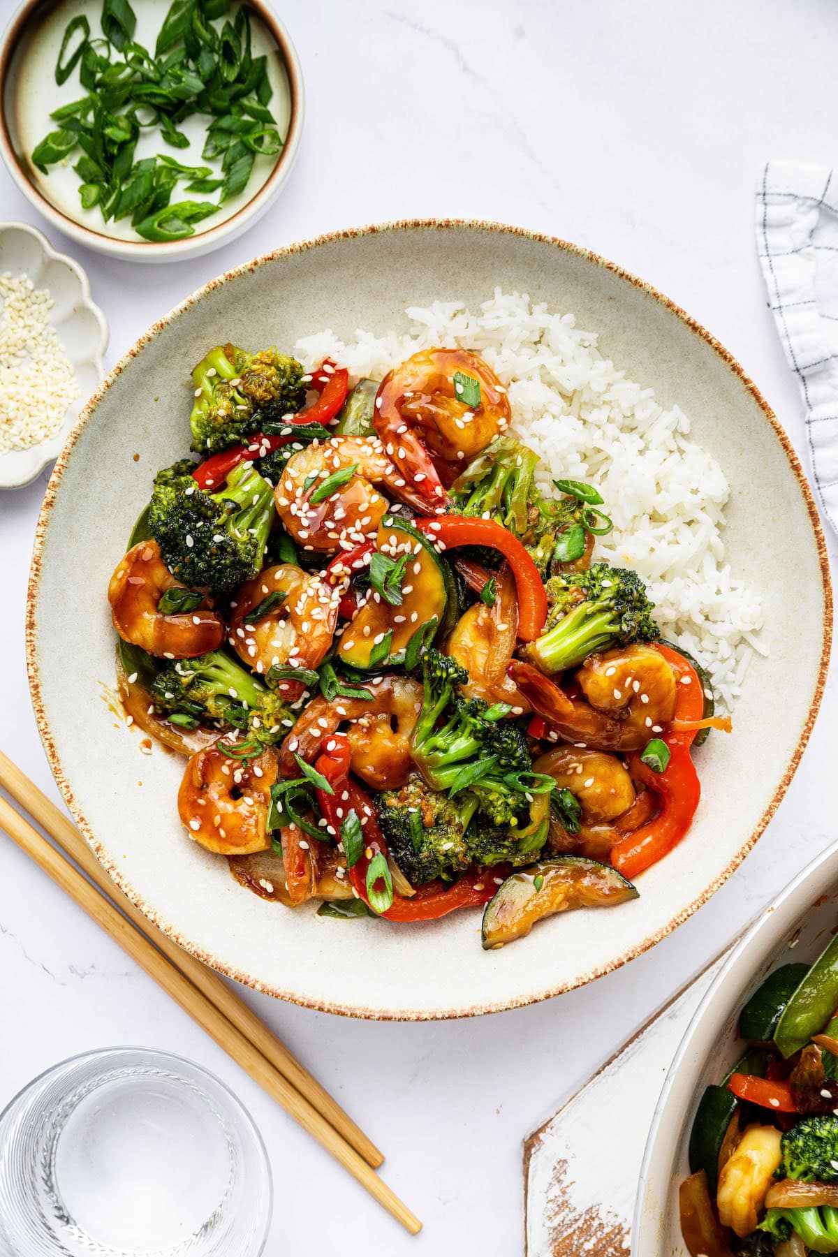 A plate of shrimp stir-fry served with white rice. The dish includes shrimp, broccoli, red bell peppers, and snap peas in a sauce, topped with sesame seeds and green onions. Chopsticks are beside the plate, with small bowls of sesame seeds and chopped green onions nearby.