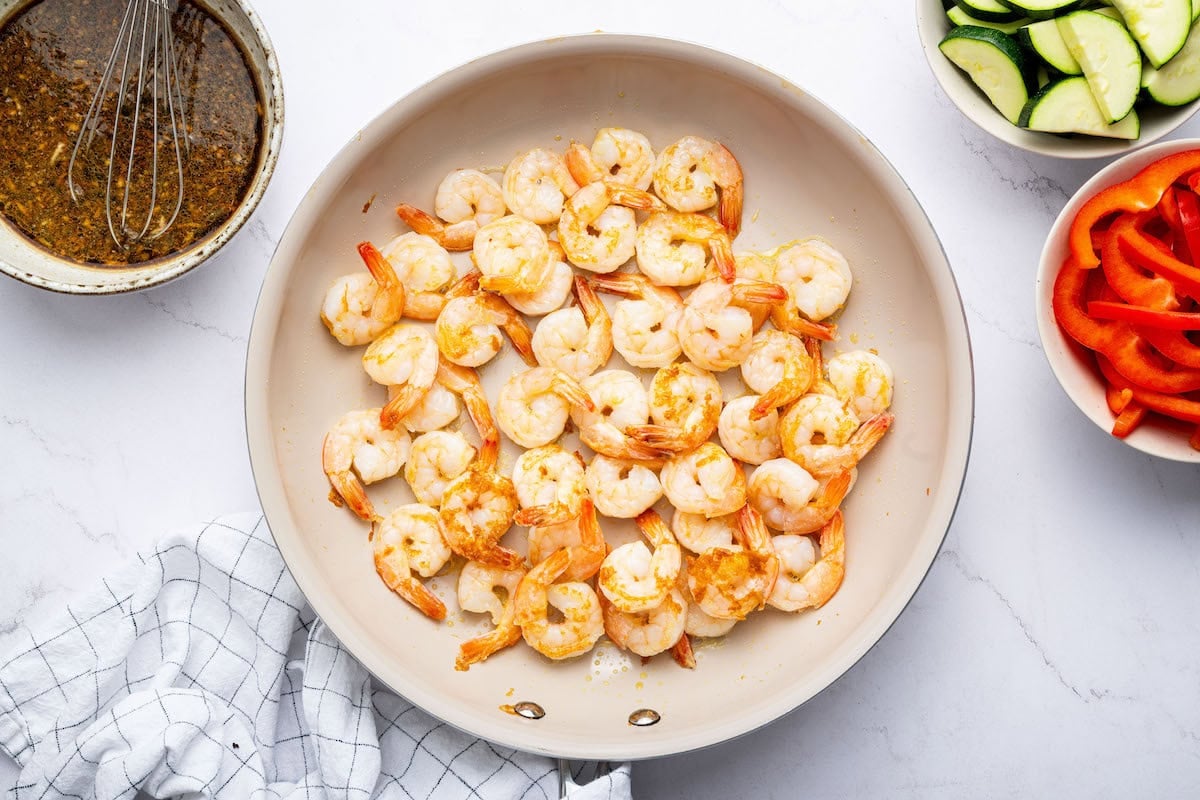 A pan of shrimp cooking, turning opaque with slight browning. To the side, there is a bowl of mixed sauce, a whisk, and bowls of sliced zucchini and red bell peppers.