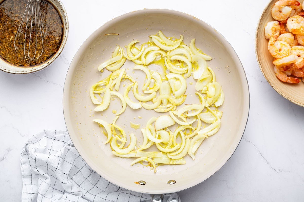 Sliced onions cooking in oil in a pan. The shrimp has been set aside in a separate bowl, and the sauce mixture is nearby with a whisk resting inside.