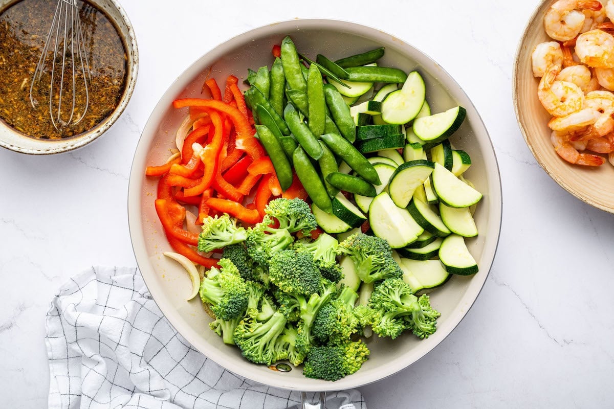 A pan filled with red bell peppers, snap peas, zucchini, and broccoli, ready to be cooked. The sauce bowl and shrimp are placed to the side.