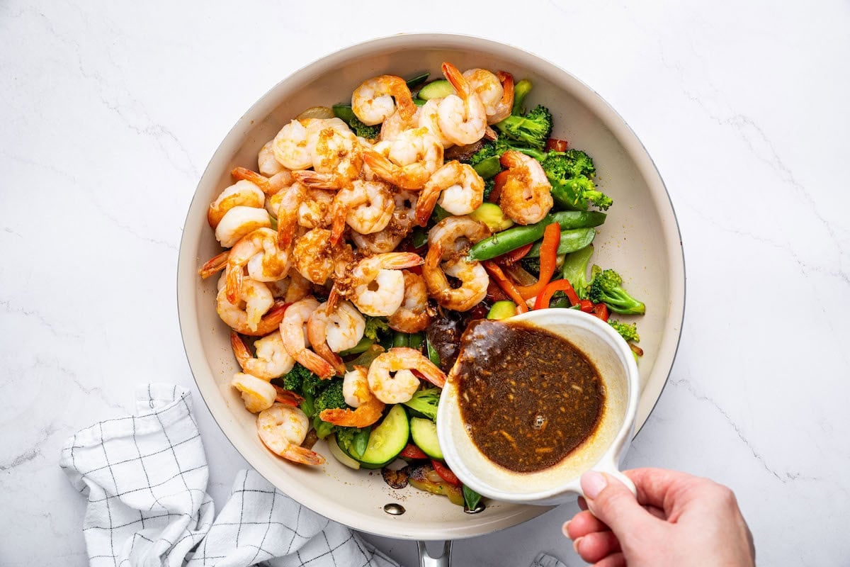 Cooked shrimp added back into the pan with the sautéed vegetables, while a hand is pouring the sauce over the mixture.