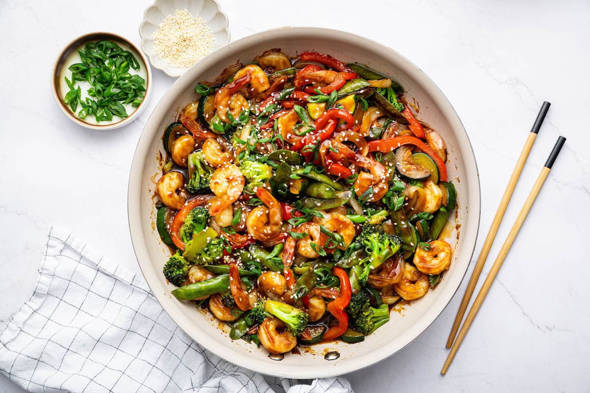 A completed shrimp stir-fry in a pan with shrimp, vegetables, and sauce mixed together. The dish is topped with sesame seeds and green onions, with a pair of chopsticks placed beside the pan.