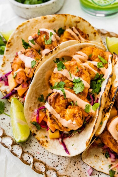 Three shrimp tacos in flour tortillas folded up on a serving platter. The tacos are topped with a mango cabbage slaw and a drizzle of spicy crema.
