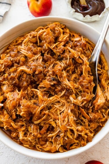 Shredded apple BBQ chicken in a white bowl with a serving spoon.