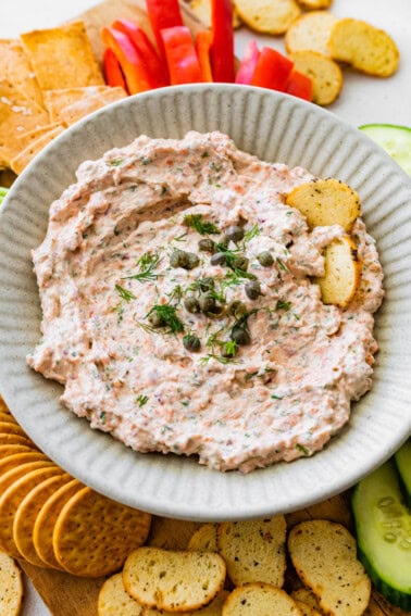 A bowl of smoked salmon dip, garnished with fresh dill and capers, served with crackers and fresh vegetables.