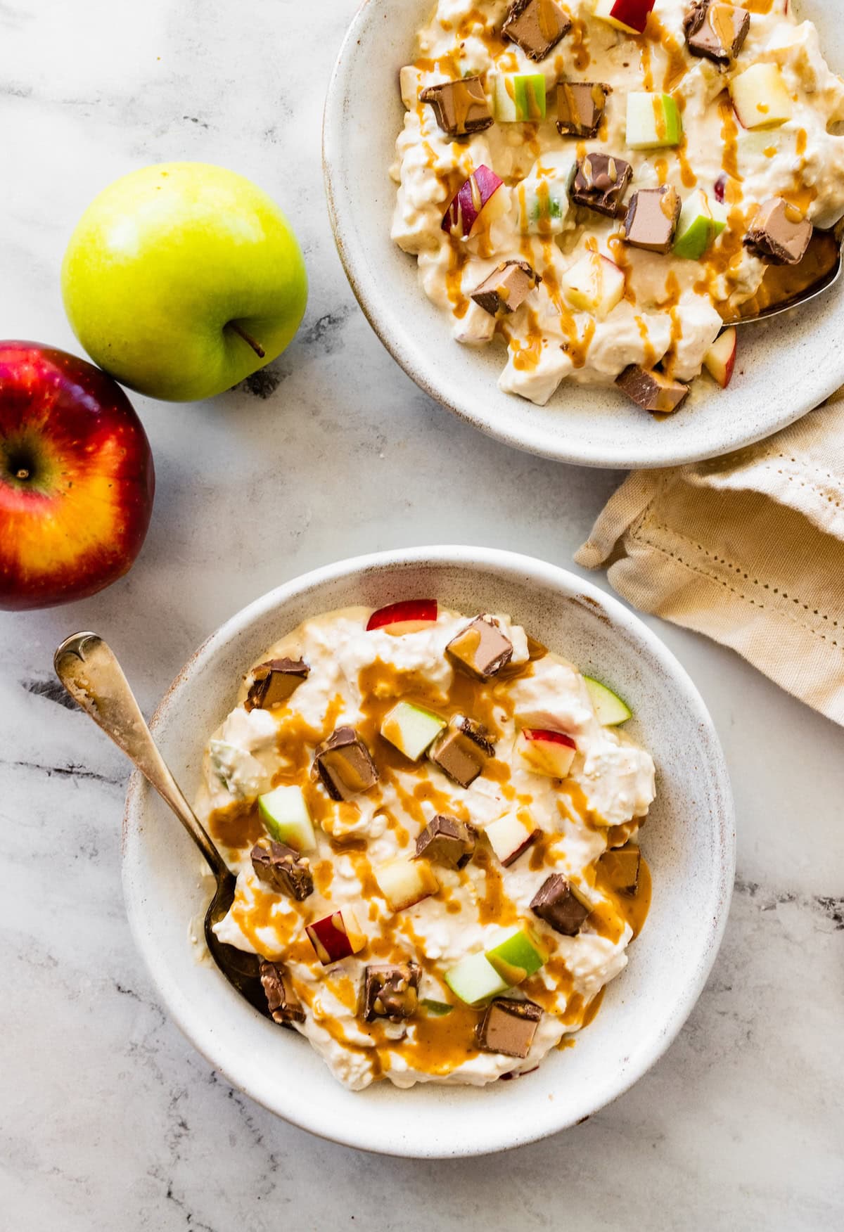 Two snickers salad bowls topped with a chopped chocolate peanut butter protein bar, apples and a drizzle of peanut butter.