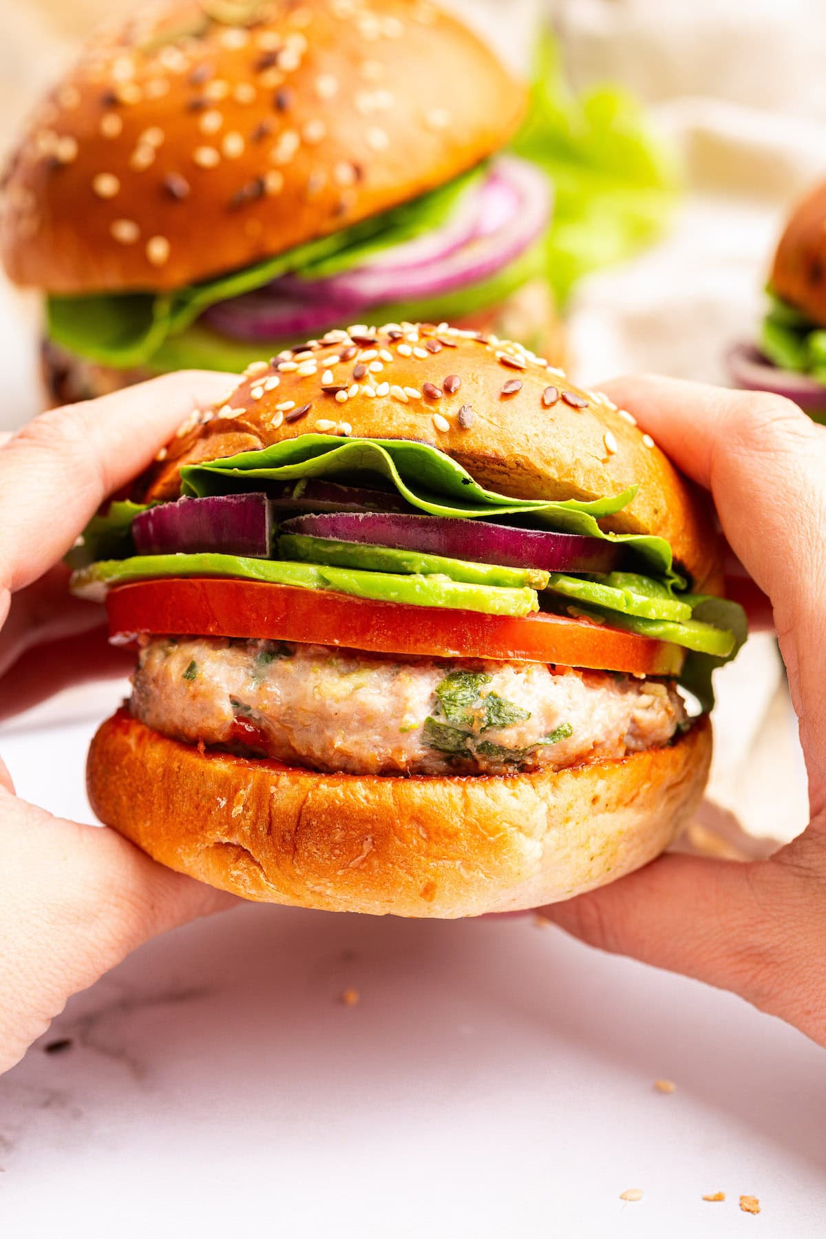 A woman's hands holding a spinach feta turkey burger served on a seeded bun with tomato, lettuce, red onion and avocado.