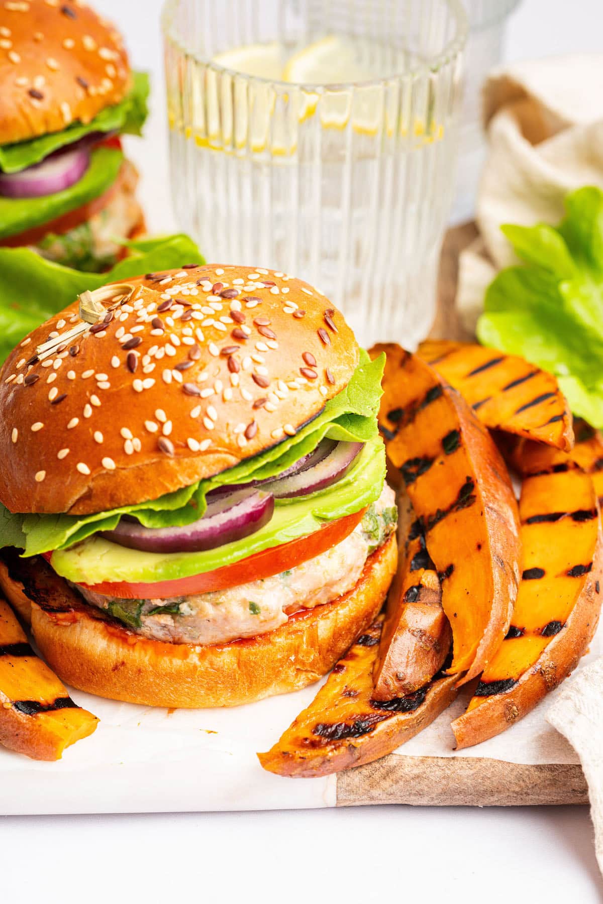 A spinach feta turkey burger served on a seeded bun with tomato, lettuce, red onion and avocado. There are grilled sweet potato wedges next to the burger and a glass of water behind it.