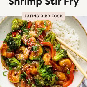 This image shows a delicious shrimp stir-fry served with white rice. The stir-fry includes vibrant vegetables like broccoli, red bell peppers, and zucchini, all coated in a glossy, savory sauce. The dish is garnished with sesame seeds and sliced green onions, adding texture and freshness. Chopsticks are placed on the side, ready for use.